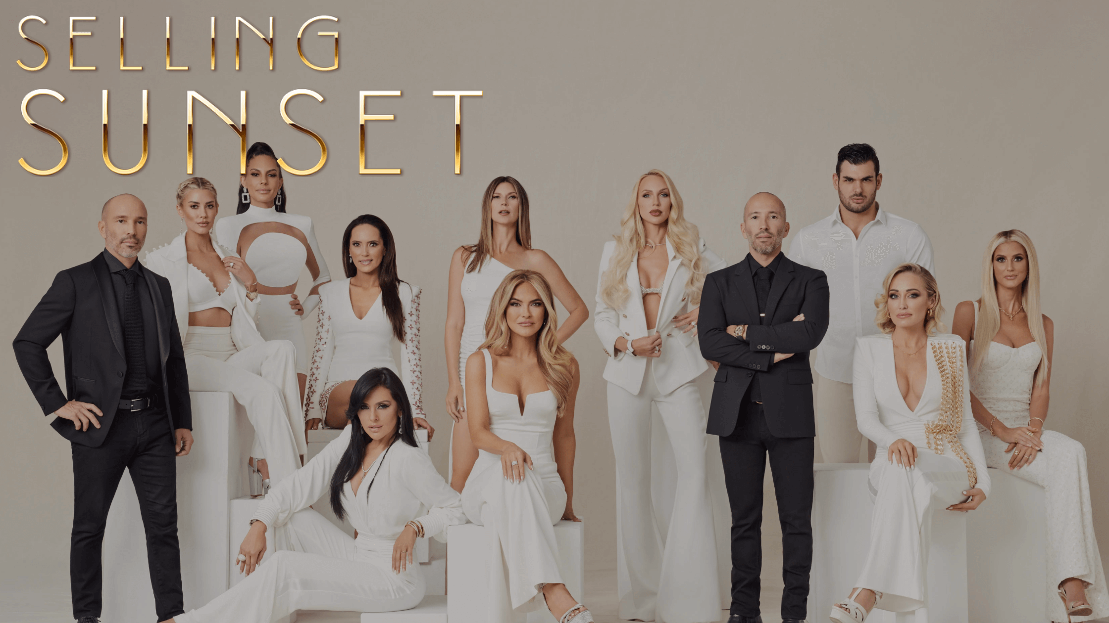 This image features a promotional group photo for the TV show "Selling Sunset". There are eleven individuals, presumably real estate agents, stylishly dressed in coordinated white and black attire, posing against a neutral beige background. The title "SELLING SUNSET" is displayed prominently at the top in large, elegant golden letters that stand out against the background. The individuals are arranged in a pyramid formation with some seated and others standing, giving a sense of hierarchy and team dynamics. The lighting is soft and professional, highlighting the polished aesthetics of the individuals and their fashionable outfits, which range from suits to elegant dresses, indicating the show's focus on luxury and glamour.